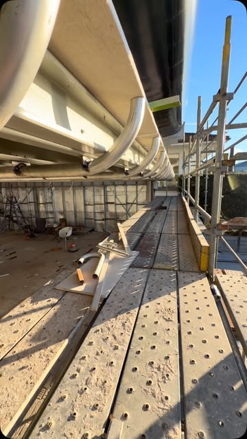 Structural steel beams installed beneath a roofline with scaffold planking running alongside.