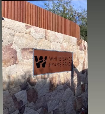 Fabricated metal entry sign reading White Sands Hyams Beach mounted on a sandstone feature wall.